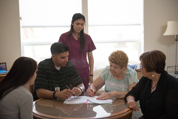 Image of a woman signing her advance directive document at the kitchen table surrounded by family 